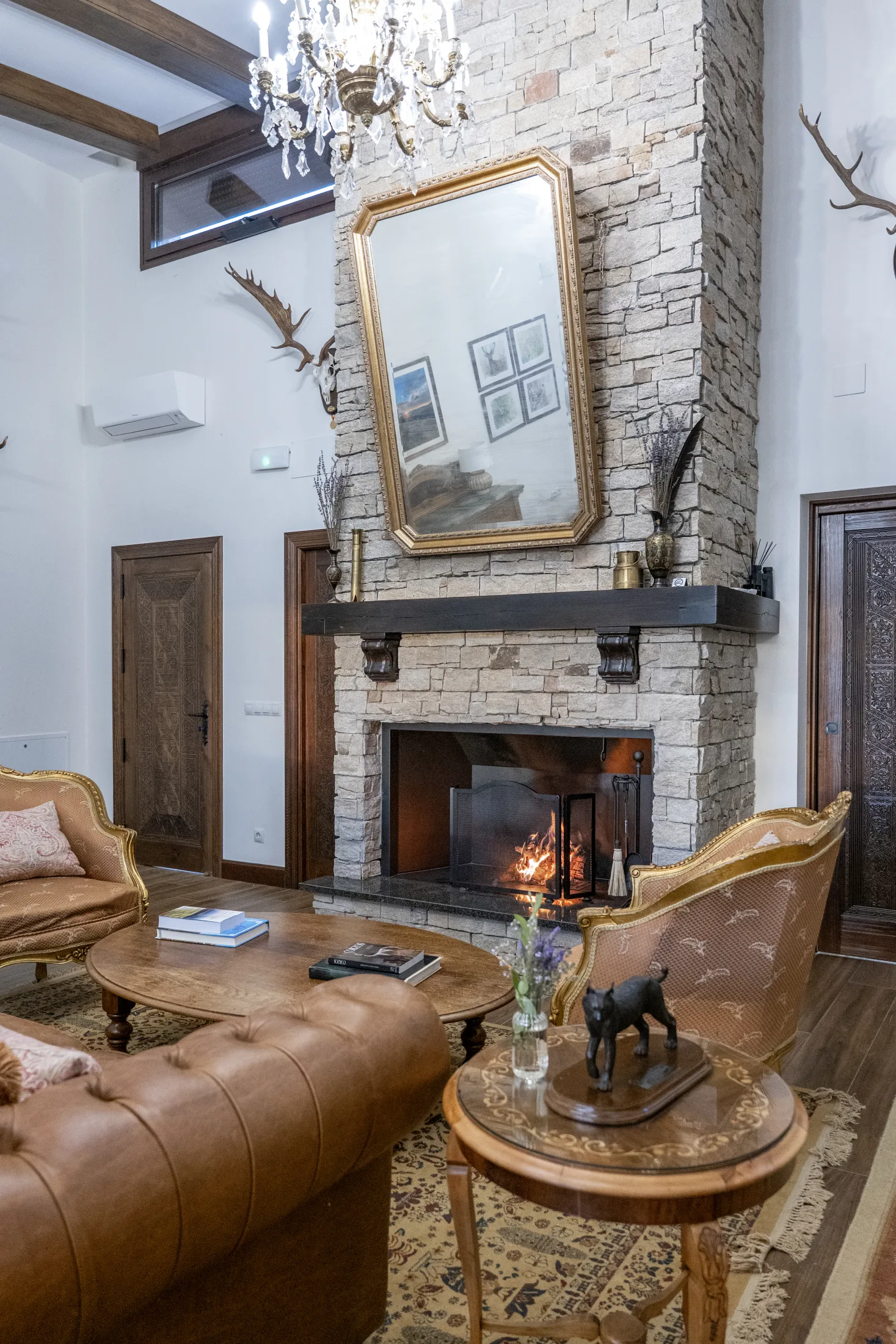 The lodge fireplace, a stone chimney with a gilt mirror, antler mounts and a lit fire below