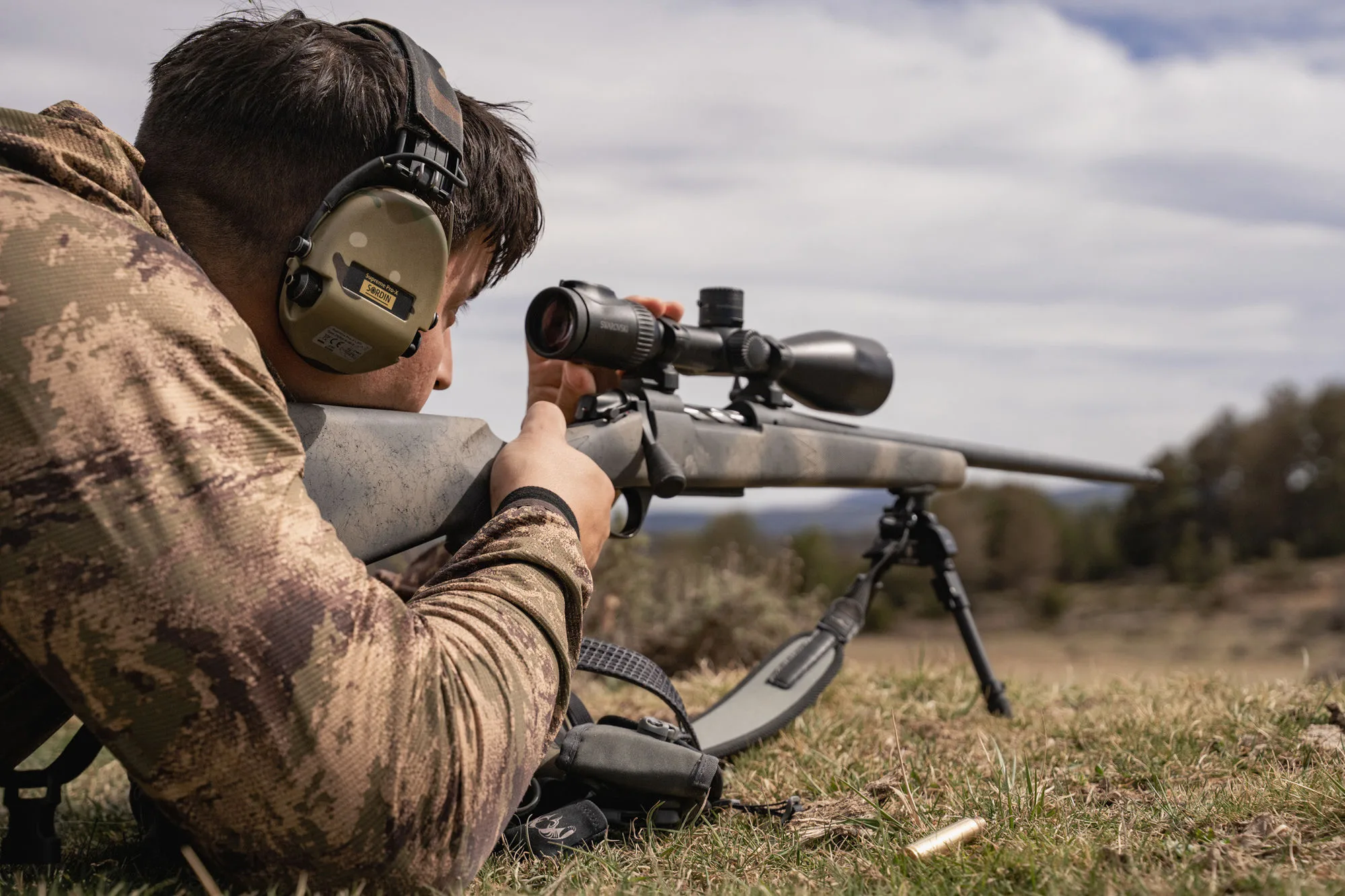 A hunter prone behind a scoped rifle on its bipod, glassing the sierra